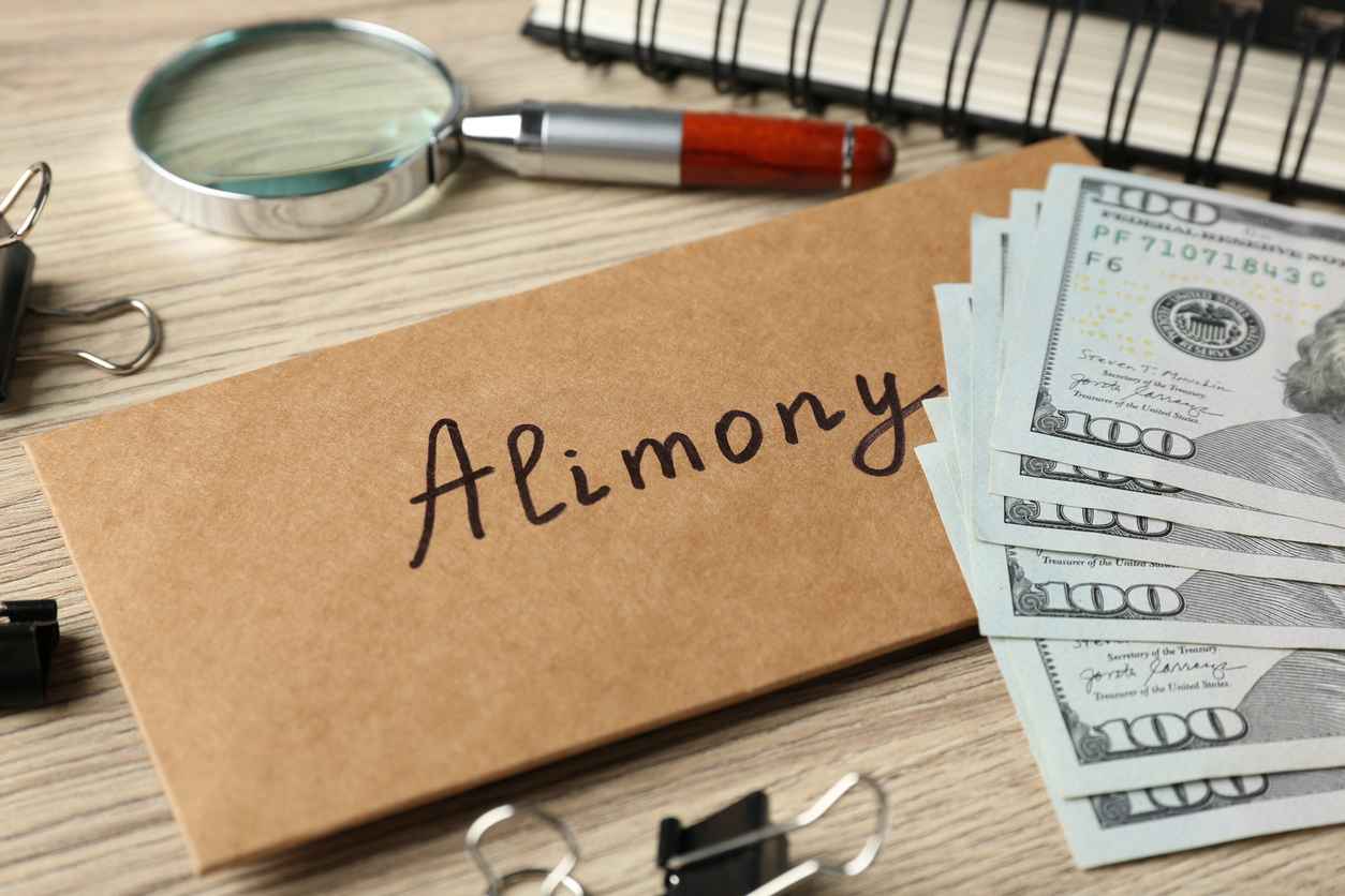 Envelope with word Alimony, money, notebook, binder clips and magnifying glass on wooden table, closeup